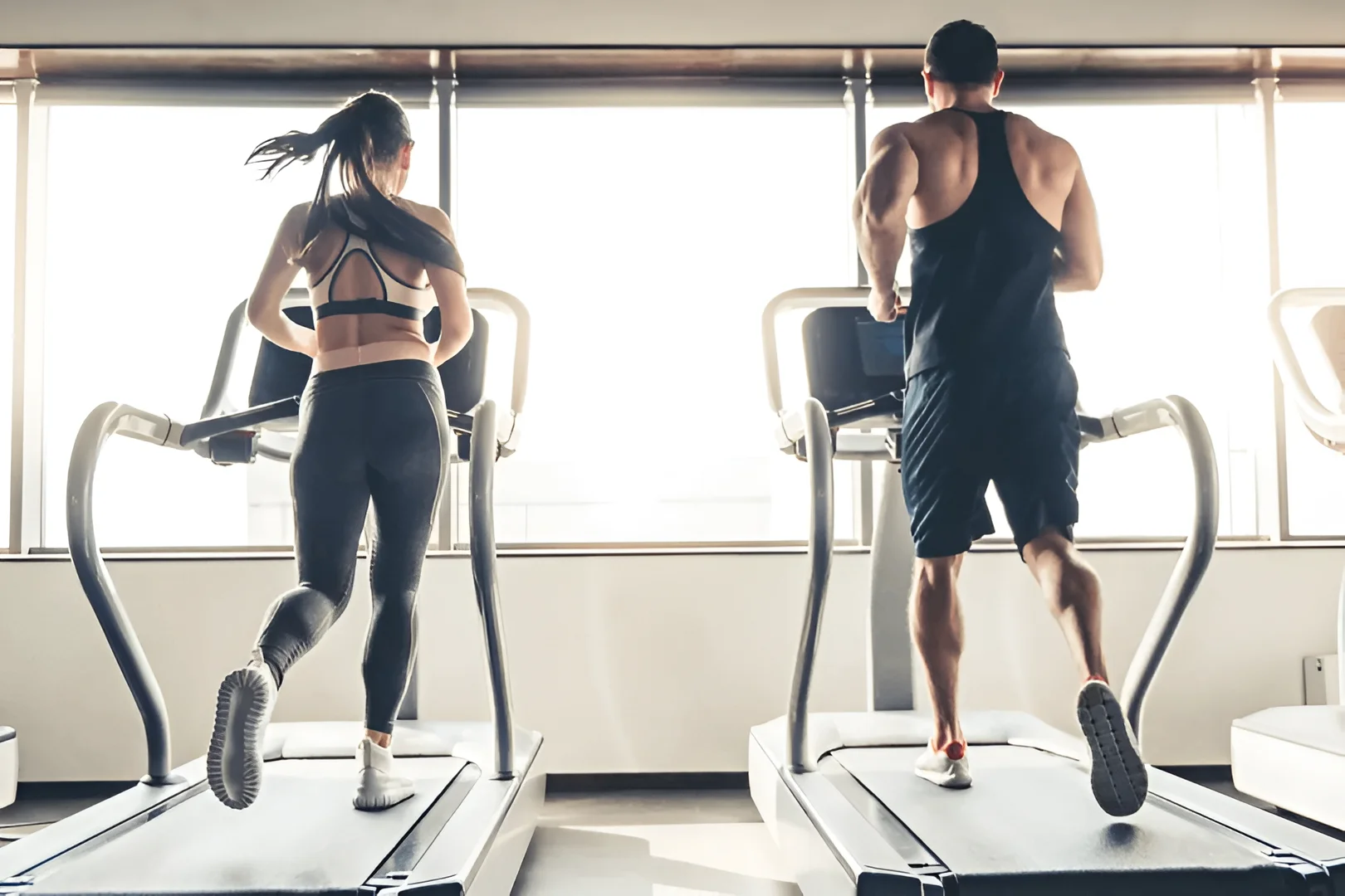 Two people are running on a treadmill in front of a window.